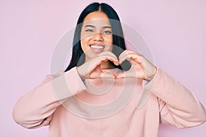 Young beautiful asian girl wearing casual pink sweater smiling in love doing heart symbol shape with hands