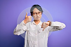 Young beautiful asian girl wearing casual shirt and glasses standing over purple background Doing thumbs up and down, disagreement