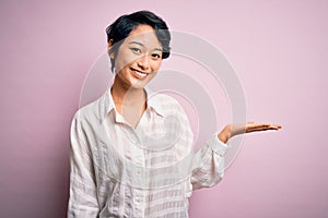 Young beautiful asian girl wearing casual shirt standing over isolated pink background smiling cheerful presenting and pointing
