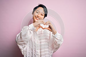 Young beautiful asian girl wearing casual shirt standing over isolated pink background smiling in love doing heart symbol shape