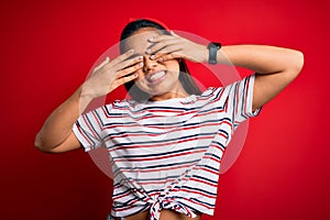Young beautiful asian girl wearing casual striped t-shirt over isolated red background covering eyes with hands smiling cheerful