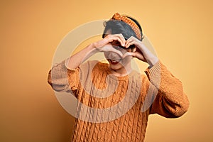 Young beautiful asian girl wearing casual sweater and diadem standing over yellow background Doing heart shape with hand and