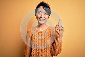 Young beautiful asian girl wearing casual sweater and diadem standing over yellow background showing and pointing up with finger