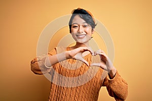 Young beautiful asian girl wearing casual sweater and diadem standing over yellow background smiling in love doing heart symbol