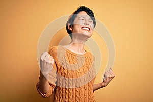 Young beautiful asian girl wearing casual sweater and diadem standing over yellow background very happy and excited doing winner