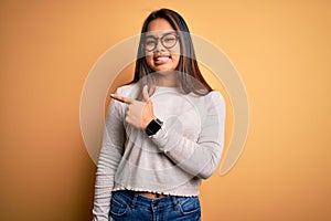 Young beautiful asian girl wearing casual sweater and glasses over yellow background cheerful with a smile of face pointing with