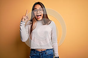 Young beautiful asian girl wearing casual sweater and glasses over yellow background pointing finger up with successful idea