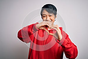 Young beautiful asian girl wearing casual sweatshirt with hoodie over white background smiling in love doing heart symbol shape