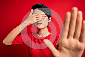 Young beautiful asian girl wearing casual t-shirt and glasses over isolated red background covering eyes with hands and doing stop