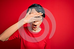 Young beautiful asian girl wearing casual t-shirt and glasses over isolated red background peeking in shock covering face and eyes