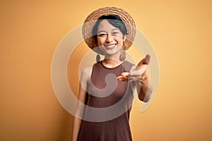 Young beautiful asian girl wearing casual t-shirt and hat over isolated yellow background smiling friendly offering handshake as