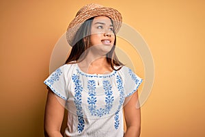 Young beautiful asian girl wearing casual t-shirt and hat standing over yellow background looking away to side with smile on face,