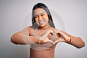 Young beautiful asian girl wearing casual turtleneck sweater standing over white background smiling in love showing heart symbol