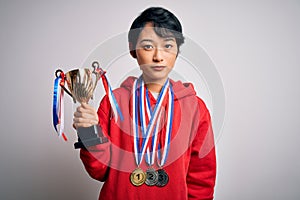 Young beautiful asian girl winner holding trophy wearing medals over white background with a confident expression on smart face