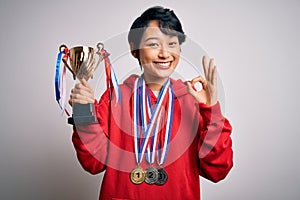 Young beautiful asian girl winner holding trophy wearing medals over white background doing ok sign with fingers, excellent symbol