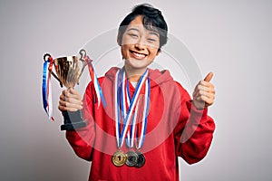 Young beautiful asian girl winner holding trophy wearing medals over white background happy with big smile doing ok sign, thumb up