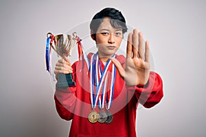 Young beautiful asian girl winner holding trophy wearing medals over white background with open hand doing stop sign with serious