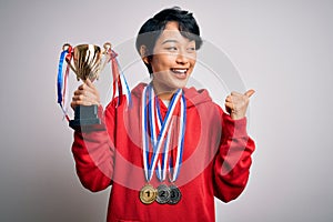 Young beautiful asian girl winner holding trophy wearing medals over white background pointing and showing with thumb up to the