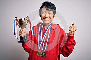 Young beautiful asian girl winner holding trophy wearing medals over white background screaming proud and celebrating victory and