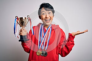 Young beautiful asian girl winner holding trophy wearing medals over white background very happy and excited, winner expression
