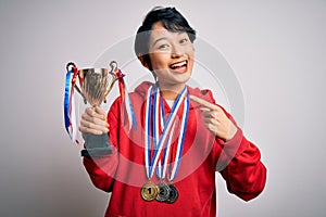 Young beautiful asian girl winner holding trophy wearing medals over white background very happy pointing with hand and finger