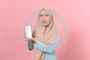 Young beautiful Asian Muslim woman wearing Islamic hijab holding a smartphone showing screen scared in shock with a surprised face