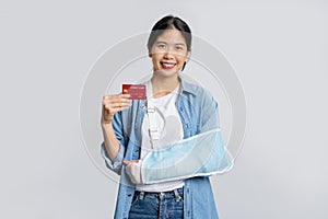 Young beautiful asian office worker in cast due to broken arm in accident while holding credit card isolated on white background.