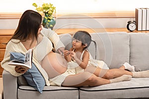 Young beautiful Asian pregnant women reading the book and her toddler daughter laying down having ear on mother tummy. Little girl