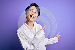 Young beautiful asian scientist girl wearing coat and glasses over purple background smiling and looking at the camera pointing