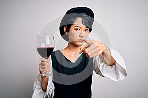 Young beautiful asian sommelier girl drinking glass of red wine over isolated white background pointing with finger to the camera