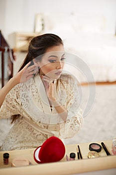 A young beautiful Asian woman in a beige lace dressing gown put on morning makeup and wears earrings . shallow depth of focus.