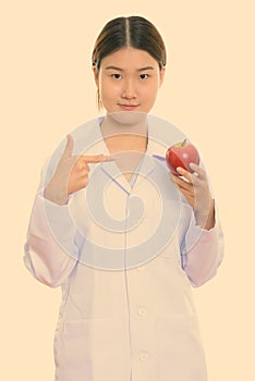 Young beautiful Asian woman doctor holding and pointing at red apple while looking at camera