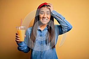 Young beautiful asian woman drinking healthy glass of orange juice over yellow background stressed with hand on head, shocked with