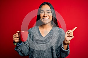 Young beautiful asian woman drinking mug of coffee standing over isolated red background very happy pointing with hand and finger