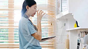 Young beautiful asian woman drinking water while holding digital tablet and standing by window in kitchen background