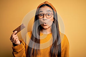 Young beautiful asian woman eating healthy protein bar over isolated yellow background scared in shock with a surprise face,