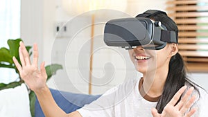 Young beautiful asian woman exciting in VR headset looking up and trying to touch objects in virtual reality at home living room,