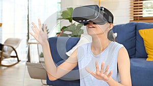 Young beautiful asian woman exciting in VR headset looking up and trying to touch objects in virtual reality at home living room,