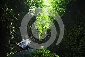 Young beautiful Asian woman practicing Yoga posing sitting in lotus position meditating over a stone in a stunning natural landsca