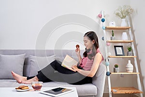 Young beautiful asian woman sitting on couch reading a book enjoying her tea in living room at home