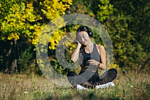 Young beautiful Asian woman sitting in lotus position in the park, relaxing after fitness and running, listening to music on