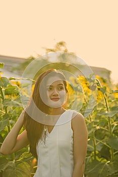 Young beautiful Asian woman thinking while fixing hair in the field of blooming sunflowers