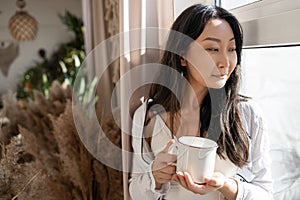 Young beautiful Asian woman in underwear drinking coffee while standing near the window in the morning.