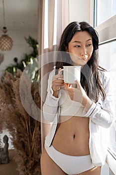 Young beautiful Asian woman in underwear drinking coffee while standing near the window in the morning.