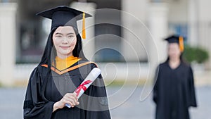 A young beautiful Asian woman university graduate in graduation gown and mortarboard holds a degree certificate stands in front of