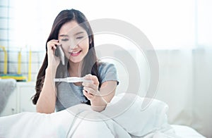 Young beautiful asian woman using smart mobile phone hand holding positive pregnancy test with smile and happy sitting on bed in