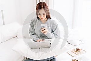 Young beautiful Asian woman in warm knitted clothes with cup of coffee and working on a laptop sitting on the bed in the house.