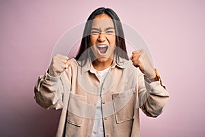 Young beautiful asian woman wearing casual shirt standing over pink background angry and mad raising fists frustrated and furious