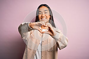 Young beautiful asian woman wearing casual shirt standing over pink background smiling in love doing heart symbol shape with hands