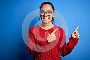 Young beautiful asian woman wearing casual sweater and glasses over blue background smiling and looking at the camera pointing